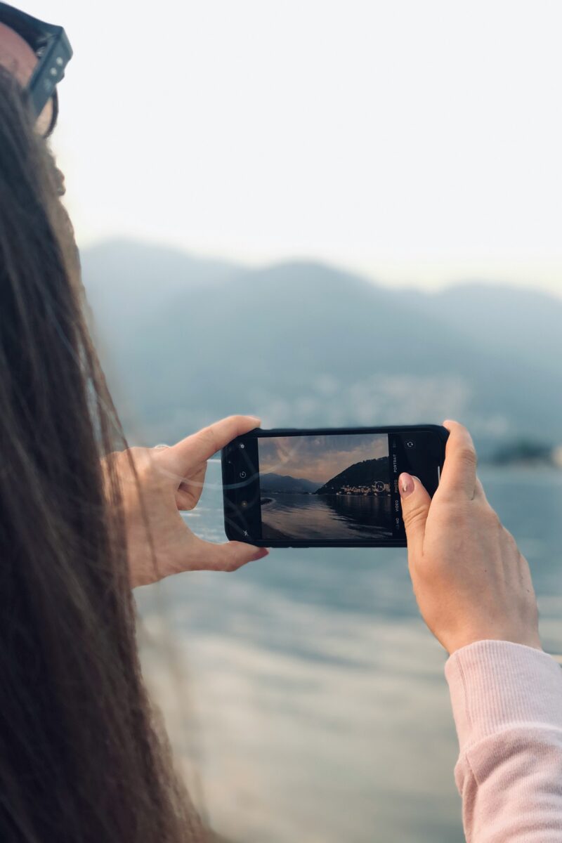 Personne tenant son téléphone intelligent pour photographier un lac et des montagnes.