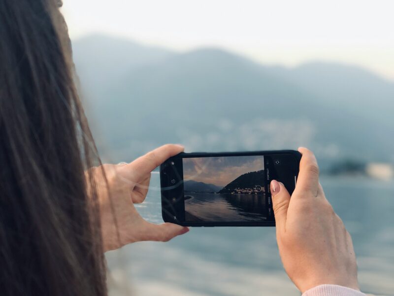 Personne tenant son téléphone intelligent pour photographier un lac et des montagnes.